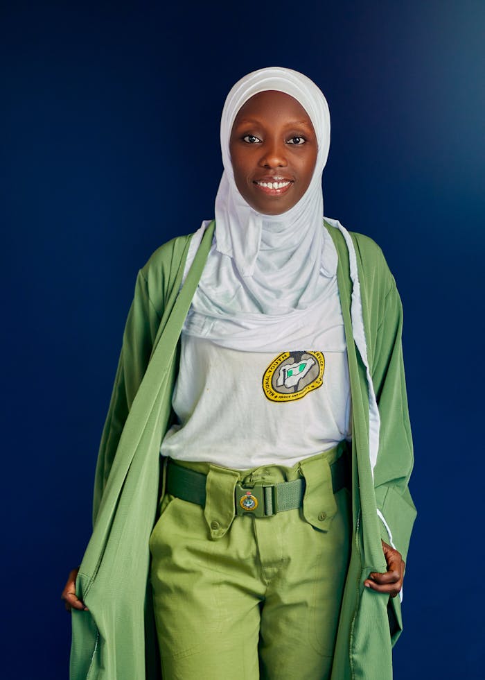 Portrait of a young woman in a green uniform and white hijab, smiling confidently.
