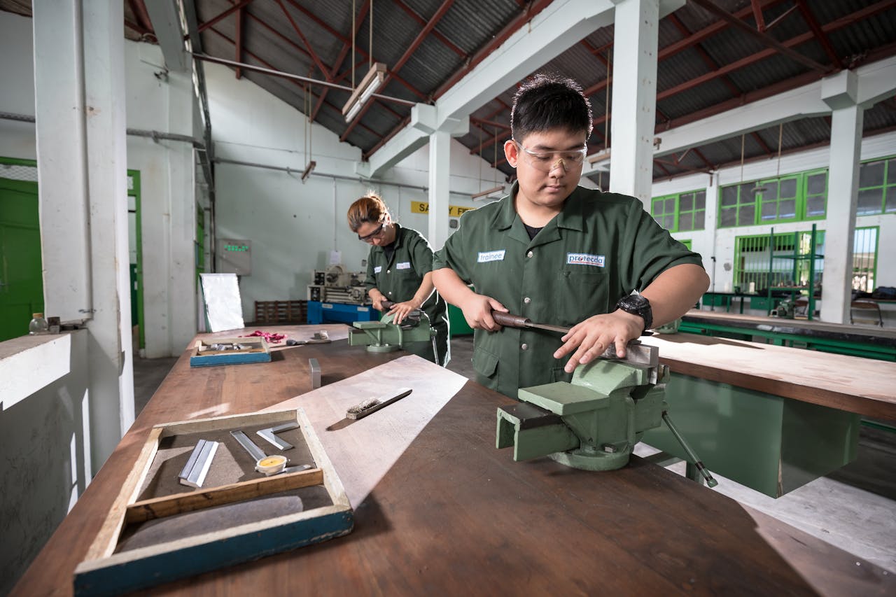 Two workers in a well-lit Indonesian workshop using clamping tools and safety gear for metalwork.